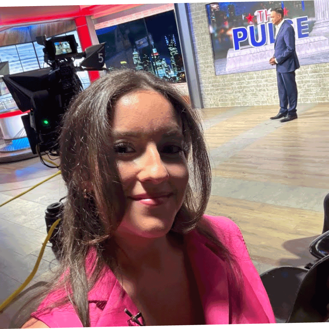 Eine Frau mit langen Haaren und pinkem Blazer macht ein Selfie in einem Fernsehstudio. Im Hintergrund steht ein männlicher Moderator auf einer Bühne neben einem großen Bildschirm mit dem Titel „THE PULSE“, und durch die Studiofenster sind Lichter der Stadt zu sehen.