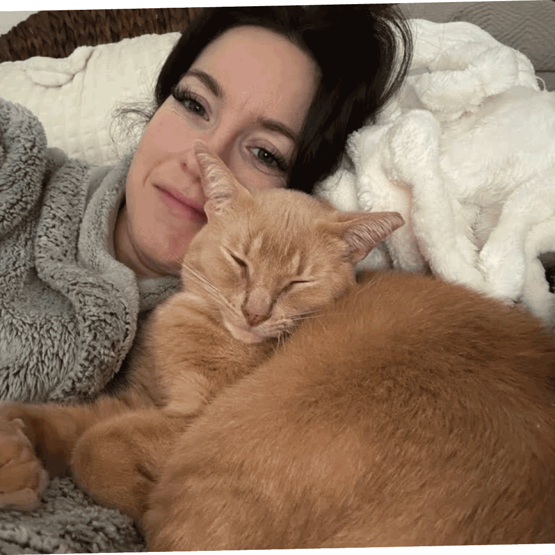 Eine Frau liegt mit einer gemütlichen Decke auf einem Sofa, lächelt sanft und kuschelt mit einer schlafenden orangefarbenen Katze. Die Katze liegt eng an ihrer Seite; beide wirken entspannt.