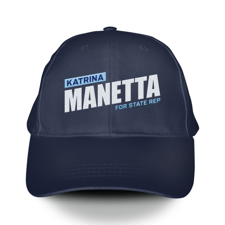 کڵاوێکی بیسبۆڵی شینی تاریک کە ناوی "Katrina Manetta" بە پیتی سپی تۆخ لەسەریەتی، لەگەڵ دەستەواژەی "FOR STATE REP" بە پیتی بچووکتر لە خوارەوە. کڵاوەکە شێوەیەکی ڕێکخراو و لێواری خەم‌دار هەیە.