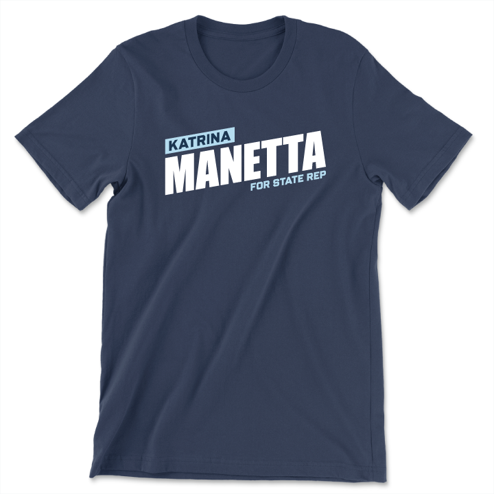 تی‌شێرتێکی شینی تاریک کە دەقی 'Katrina Manetta for State Rep' بە پیتی سپی و شینی کاڵی تۆخ لەسەریەتی. تی‌شێرتەکە ملی گەرد و قۆڵی کورت هەیە.